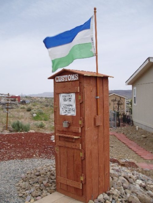 A wooden customs booth with a flag on top, featuring a sign and surrounded by gravel and rocks.