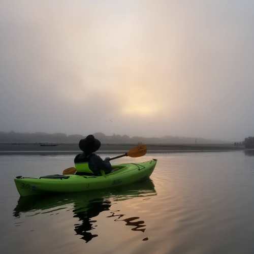 A person in a kayak paddles through a misty lake at sunrise, surrounded by calm waters and soft fog.