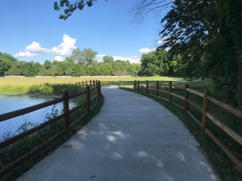 A paved path curves alongside a pond, bordered by wooden fencing and surrounded by lush greenery under a blue sky.