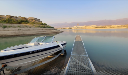 A boat is docked at a calm lake with sandy shores and distant mountains under a clear sky.