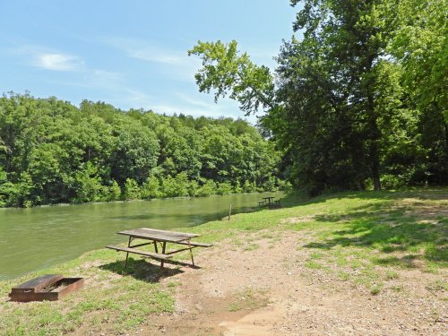 A serene riverside scene with picnic tables, lush green trees, and calm water under a clear blue sky.