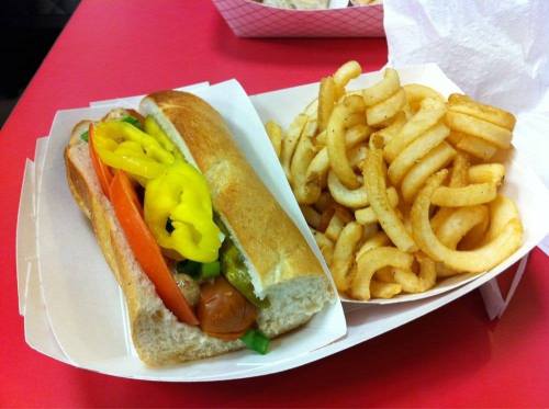 A hot dog in a bun topped with tomatoes, peppers, and onions, served with a side of curly fries on a red table.