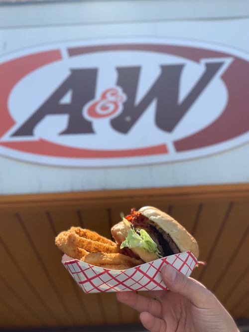 A hand holding a burger and fries in a red and white basket, with an A&W sign in the background.