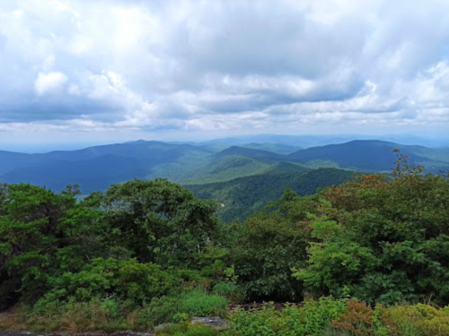 A panoramic view of rolling green mountains under a cloudy sky, with lush trees in the foreground.