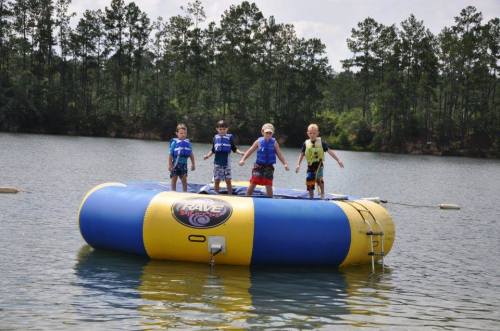 Four children in life jackets stand on a large inflatable water trampoline in a lake, surrounded by trees.