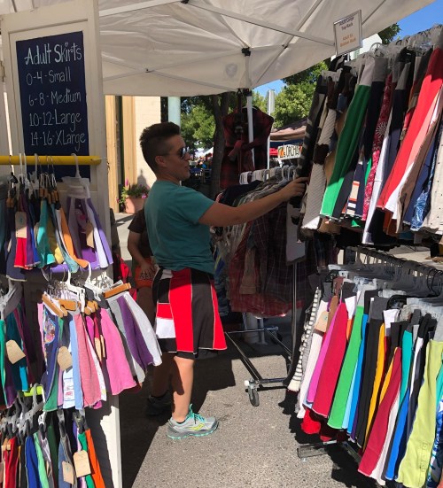 A person browses colorful adult skirts at a market stall under a canopy on a sunny day.