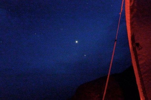 Night sky with stars and two bright celestial objects, illuminated by a faint red light from a nearby structure.