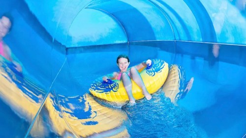 A child smiles while riding a yellow inflatable tube down a blue water slide.