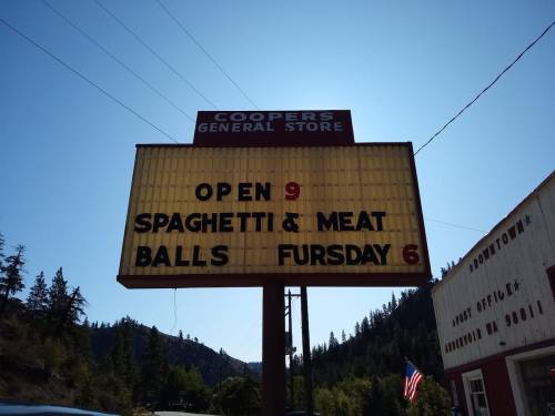 Sign for Cooper's General Store: "Open 9. Spaghetti & Meatballs Thursday 6." Surrounded by trees and blue sky.