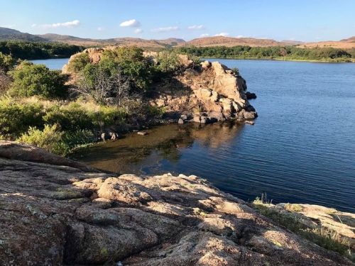 A rocky shoreline with lush greenery and calm water under a clear blue sky, surrounded by distant hills.