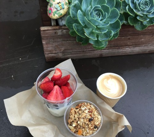 A wooden tray with a cup of yogurt topped with strawberries, a bowl of granola, and a small cup of cream, alongside succulents.
