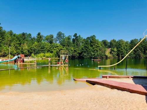 A sunny lakeside scene with a sandy beach, water slides, and a floating playground surrounded by trees.