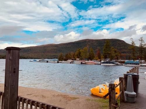 A serene lake view with boats docked, surrounded by trees and mountains under a partly cloudy sky.