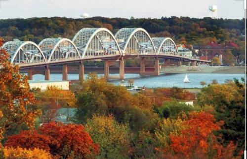 A scenic view of a bridge over a river, surrounded by autumn foliage and a small sailboat in the water.
