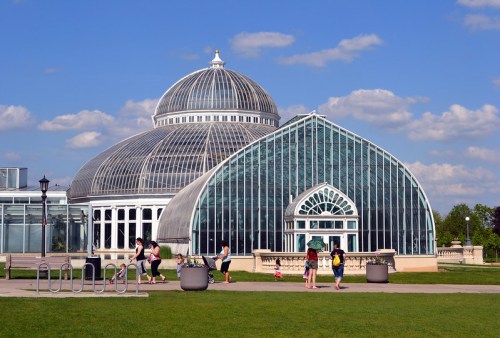 A large glass conservatory with a dome, surrounded by green grass and people walking nearby under a blue sky.