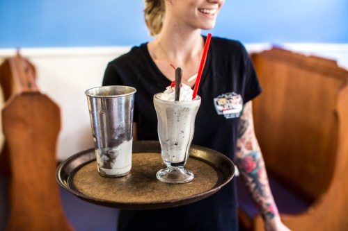 A server holds a tray with a milkshake in a glass and a metal cup, smiling in a casual dining setting.