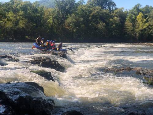 A group of people in a raft navigating a rocky river with rapids, surrounded by trees and a clear blue sky.