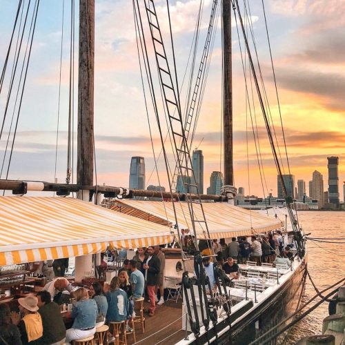 A bustling waterfront scene with a sailboat, people dining under striped awnings, and a city skyline in the background.