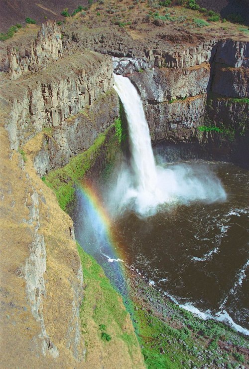 A waterfall cascades into a rocky canyon, creating mist and a rainbow in the sunlight. Lush greenery surrounds the scene.