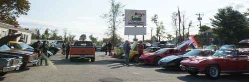 A car show at Billy's Marketplace featuring various classic and sports cars, with people admiring the vehicles.