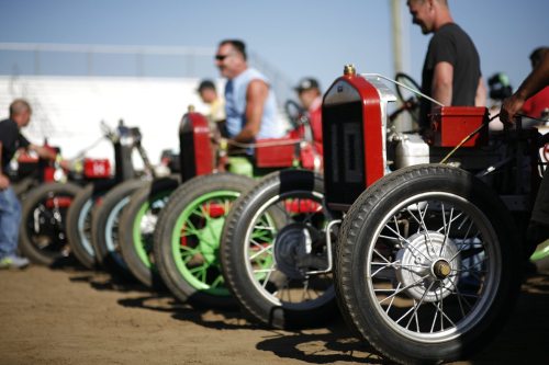 A lineup of vintage race cars with large wheels, set on a dirt track, with spectators in the background.