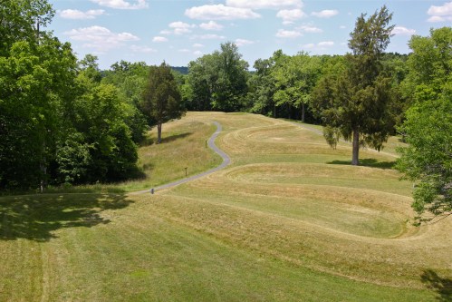 A winding path through a grassy landscape surrounded by trees under a blue sky with fluffy clouds.