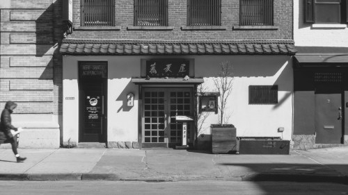 A black and white photo of a storefront with a traditional roof, featuring a door and a small tree outside.