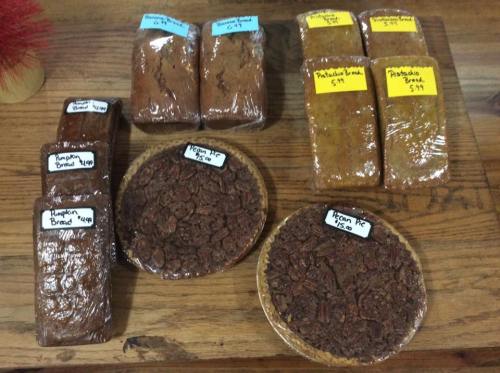 A wooden table displays various wrapped baked goods, including pumpkin bread, pecan pie, and pistachio bread, with price tags.