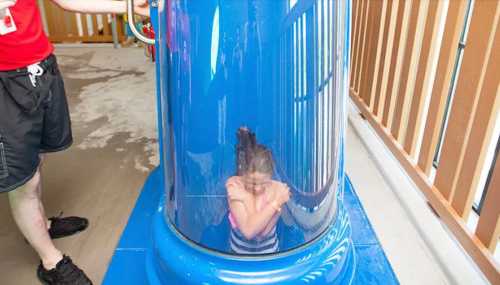 A child sits inside a clear water slide tube, preparing to go down, while an adult stands nearby.