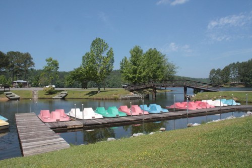 Colorful paddle boats lined up on a dock by a serene lake, with a wooden bridge and lush greenery in the background.