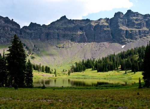 A serene mountain landscape featuring a calm lake surrounded by lush greenery and towering rocky cliffs.