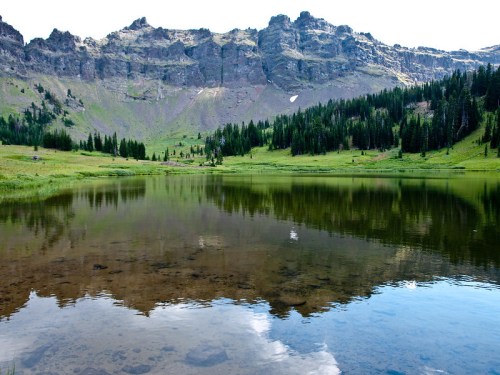 A serene lake reflecting mountains and trees, surrounded by lush green meadows under a clear blue sky.