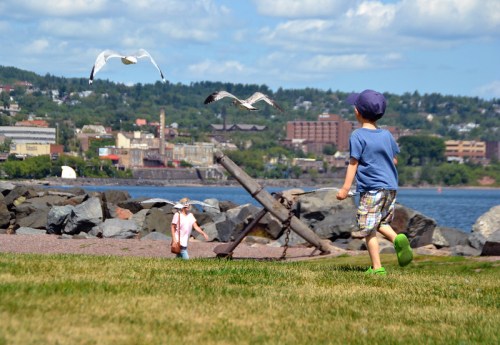 Two children play near the water, with seagulls flying overhead and a scenic view of the town in the background.