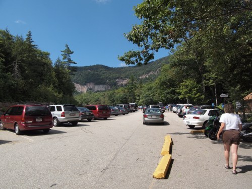 A busy parking lot surrounded by trees and mountains, with cars parked on both sides and a person walking.