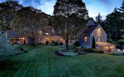 A spacious house surrounded by trees and a well-maintained lawn, illuminated at dusk with warm lights.