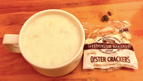 A cup of creamy soup next to a packet of Westminster Bakers oyster crackers on a wooden table.