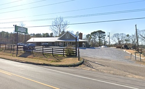 A gravel parking lot in front of a building with a sign, surrounded by trees and power lines on a clear day.