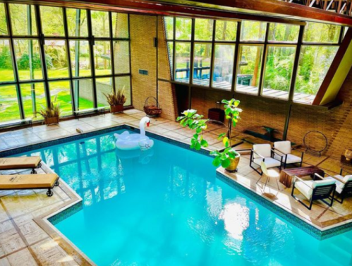 A spacious indoor pool surrounded by large windows, lounge chairs, and a swan float, with greenery outside.