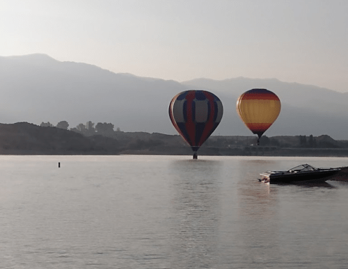 Two hot air balloons float over a calm lake, with mountains in the background and a boat nearby.