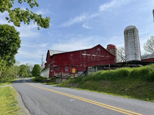 A red barn with silos beside a quiet country road, surrounded by green trees and grass under a clear blue sky.