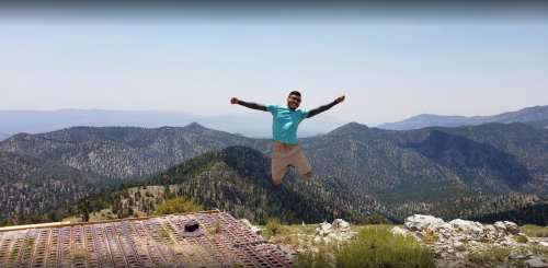 A person jumps joyfully on a mountain peak, surrounded by lush green hills and a clear blue sky.
