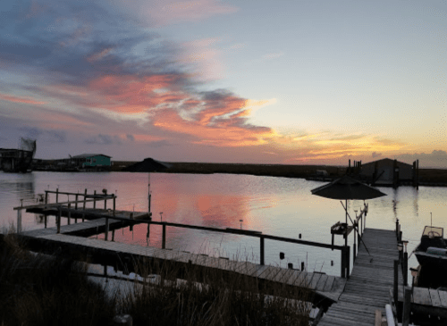 A serene sunset over a calm waterway, with colorful clouds reflecting on the surface and a wooden dock in the foreground.