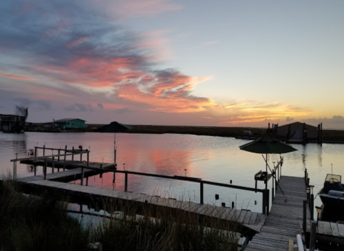 A serene sunset over a calm waterway, with colorful clouds reflecting on the surface and a wooden dock in the foreground.