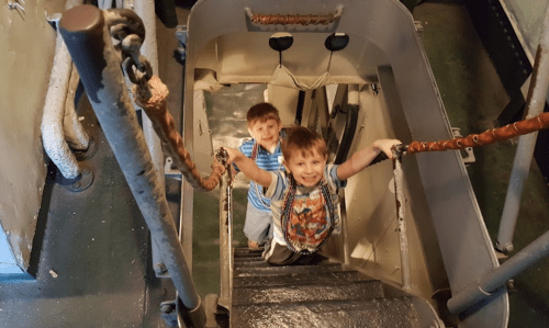 Two young boys smiling and climbing up a staircase on a ship, with metal railings and a narrow passageway.