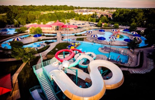 Aerial view of a water park featuring slides, pools, and colorful umbrellas surrounded by green trees.