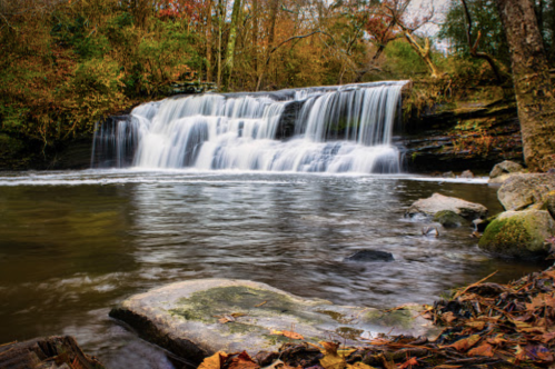 A serene waterfall cascades over rocks into a calm pool, surrounded by autumn foliage and trees.