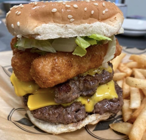 A towering burger with two beef patties, melted cheese, lettuce, onions, and crispy onion rings, served with fries.