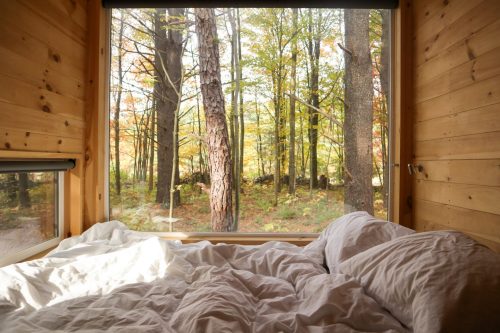 Cozy bed with rumpled white sheets, framed by a large window overlooking a serene forest in autumn.