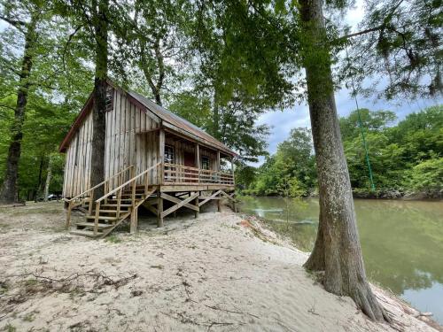 A rustic wooden cabin on stilts by a river, surrounded by trees and sandy ground.