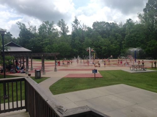 A splash pad with children playing in water, surrounded by trees and picnic areas on a cloudy day.